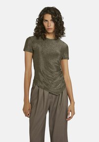 Korte mouw top in donkergroen met een glinsterende textuur, voorzien van een gedrapeerd zijdetail en een ronde halslijn. Gecombineerd met losse bruine broeken.