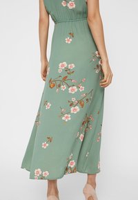 Robe maxi en tissu vert avec imprimé fleuri. Présente une taille cintrée, des manches courtes et une jupe fluide. Les accents comprennent des fleurs roses et marron.