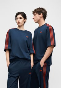 Δύο άτομα που φορούν ταυτόσημες φόρμες Adidas σε σκούρο μπλε με κόκκινες ρίγες, στέκονται δίπλα-δίπλα μπροστά σε ένα απλό φωτεινό φόντο.
