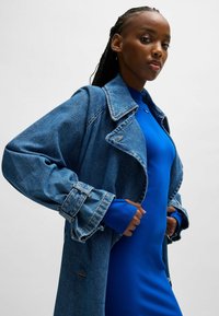 Denim trenchcoat met oversized mouwen en een kraag, gelaagd over een effen blauwe jurk. Inclusief blauwe manchetten en gestikte accenten.
