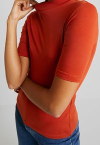 Czerwony top z krótkim rękawem i wysokim dekoltem, wykonany z gładkiego materiału. Posiada dopasowany krój z zwężającym się dołem.