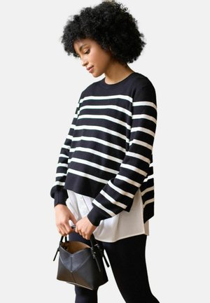 Kvinde med krøllet hår iført sort-hvidstribet sweater og hvid bluse, holder en lille sort håndtaske med begge hænder.