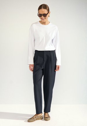 Femme portant un pantalon noir, une chemise blanche à manches longues, des lunettes de soleil et des chaussures beiges à lacets décontractées, debout sur fond blanc.