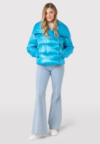 Afgekorte, glanzende blauwe pufferjack met grote kraag en drukknoopjes. Draagt over een wit shirt en gecombineerd met lichtblauwe flared jeans.