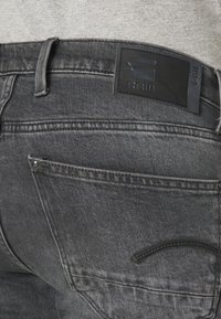Los jeans de mezclilla de un gris oscuro cuentan con un parche de cuero con la marca "RAW", costuras visibles y un diseño de bolsillo curvado sutil.
