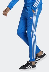 Modré tepláky s bílými bočními proužky, vyrobené z hladkého materiálu. Mají vyšívané logo na levém boku a přiléhavý střih.