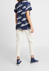 Navy blue t-shirt with large white "ellesse" logo print, paired with cream-colored trousers and navy high-top sneakers. Casual outfit composition.