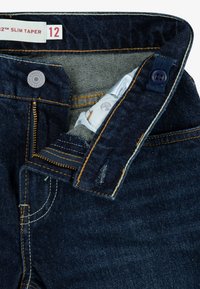 Tamno plave traperice s uskim oblikovanjem, svijetloplavim unutarnjim podstavljanjem, kontrastnim narančastim šavovima i zatvaranjem na gumbe.