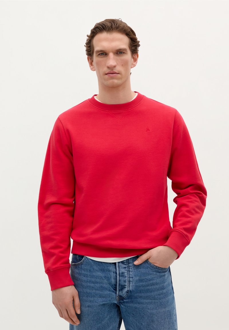 Rød sweatshirt med rund hals og ribstrikkede manchetter, lavet af blødt materiale, parret med blå jeans. Har et lille logo på brystet.