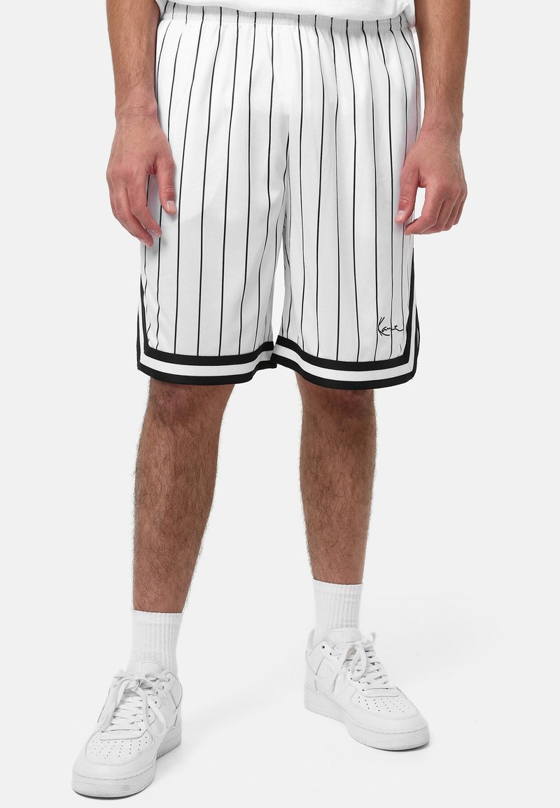 Vita sportshorts i vitt med svarta vertikala ränder, elastisk midja och svarta detaljer vid fållen. Bärs med vita sneakers och strumpor.