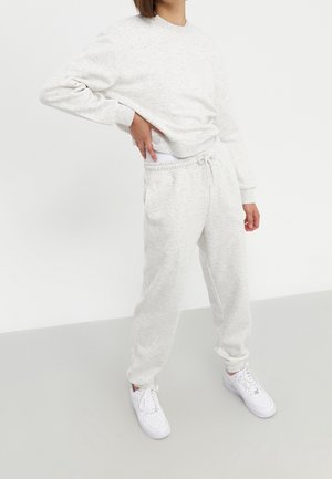 Personne portant un sweat-shirt gris clair et un pantalon de survêtement assorti avec des baskets blanches, debout avec une main sur la hanche devant un fond uni.