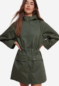 Grøn hættejakke med indsnøret talje, to frontlommer og 3/4 ærmer. Lavet af glat stof med en afslappet pasform.