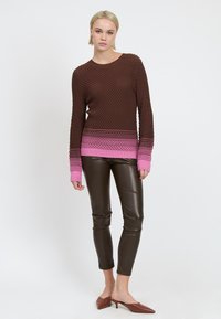 Vrouw staat in een bruine textuurtrui met roze accenten, donkere leren broek en bruine puntige kittenheels tegen een witte achtergrond.