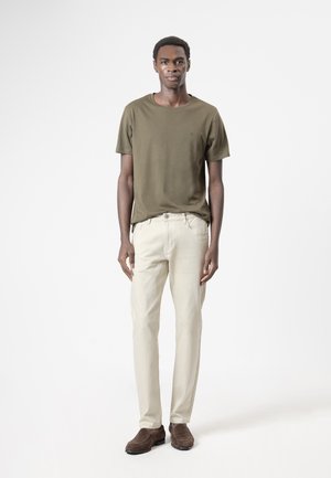 Hombre de pie, erguido, vestido con camiseta verde oliva, pantalones beige y zapatos marrones sobre un fondo blanco liso.