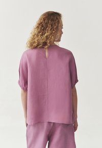 Blusa a maniche corte e morbida in lino viola chiaro con dettaglio a chiave sul retro e maniche arricciate, abbinata a pantaloni coordinati.