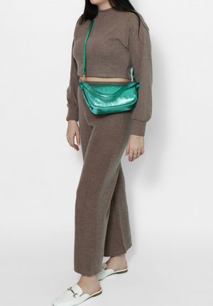 Vrouw in een bruin gebreid tweedelig outfit met witte instapschoenen en metalen groene crossbodytas voor een witte achtergrond.