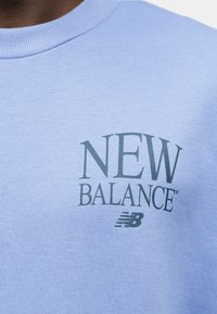 Φωτεινό μπλε φούτερ με εκτύπωση του κειμένου "NEW BALANCE" και λογότυπου "NB" σε σκούρο μπλε στο στήθος.