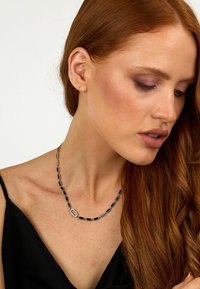 Collana rettangolare di perline argento e nera con un dettaglio ovale, indossata da un modello. Texture liscia sulla pelle nuda.