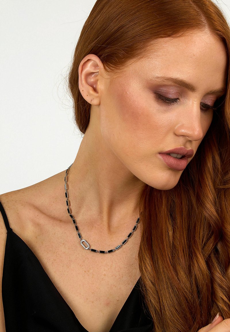 Collana rettangolare di perline argento e nera con un dettaglio ovale, indossata da un modello. Texture liscia sulla pelle nuda.