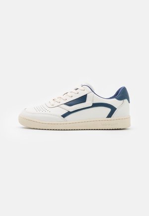 Zapatilla baja blanca con detalles en azul, puntera perforada, cierre con cordones y suela beige texturizada sobre un fondo liso.