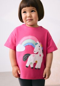 T-shirt rose à manches courtes avec un motif de licorne, des patchs, un arc-en-ciel et un nuage. La licorne est décorée de pois colorés et a une queue noire.