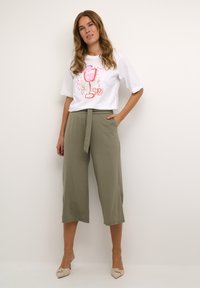 Vrouw met lang haar die een wit graphic t-shirt en olijfgroene cropped wijde broek met strikceintuur draagt, staand tegen een effen achtergrond.