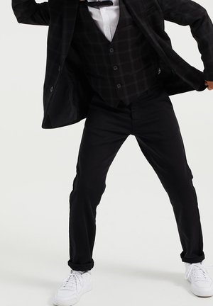 Czarna marynarka w kratę, dopasowana kamizelka, biała koszula, czarne spodnie i białe sneakersy. Marynarka noszona jest rozpięta, odsłaniając kamizelkę.