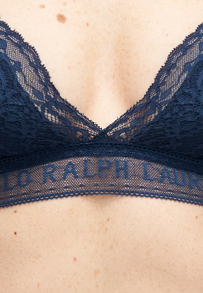 Μπλε σκούρο μπραλέτ δαντέλας με κυματιστά άκρα και ταινία με το κείμενο "Ralph Lauren", φορεμένο σε ανοιχτόχρωμο δέρμα με ορατά φακίδες.