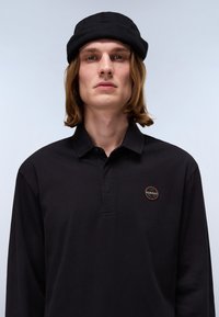 Crna dugosleeve polo majica s kragnom, s kružnim emblemom na lijevoj strani; nošena s crnom beanie kapom. Glatka tekstura.
