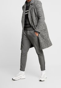 Manteau en tweed gris, pull rayé noir et gris, pantalon gris fuselé avec fermetures éclair latérales, et baskets blanches avec accents beige.