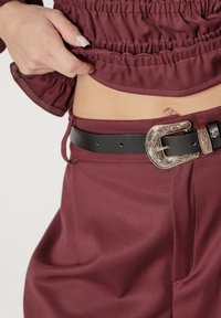 Bordo bluzka z marszczonymi detalami, zestawiona z czarnym skórzanym paskiem z ozdobną srebrną klamrą. Gładka faktura i dopasowany krój.