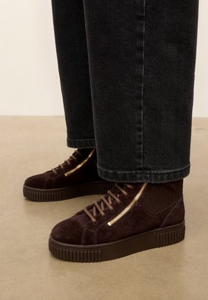 Baskets montantes en daim marron avec lacets sur le devant et fermetures éclair latérales dorées, portées avec un jean noir droit.