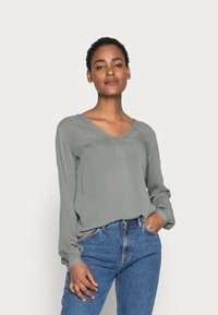 Blusa grigia a maniche lunghe con scollatura a V e una piccola tasca frontale, abbinata a jeans in denim blu. Il tessuto sembra morbido e leggermente strutturato.