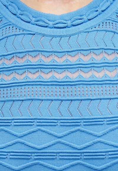 Pull en tricot bleu avec des motifs texturés variés comprenant des torsades, des zigzags et des chevrons sur une peau claire visible à travers les trous du tricot.