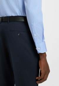 Ljusblå och vit randig skjorta med knäppningar vid ärmarna, kombinerad med mörkblå kostymbyxor med en knappfickdetalj.