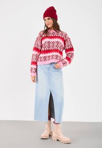 Sweter w czerwono-różowy wzór, jasnoniebieska spódnica dżinsowa z przednim rozcięciem i beżowe gumowe buty, dopełnione czerwonym beanie.