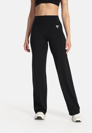 Frau trägt schwarze, hoch taillierte, weit geschnittene Hose mit weißem Logo, kombiniert mit weißen Sneakers und einem schwarzen Cropped-Top.