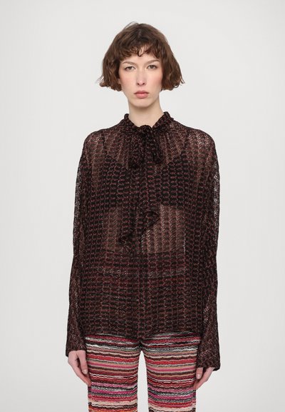 Blusa con motivo nero e rosso, con nodo al collo, tessuto trasparente, maniche lunghe e vestibilità ampia; abbinata a pantaloni a righe multicolori.