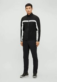 Ανδρικό μαύρο μπουφάν με φερμουάρ και άσπρες ρίγες στα μανίκια, με ψηλό γιακά. Συνδυασμένο με σκούρα παντελόνια και μαύρα αθλητικά παπούτσια.