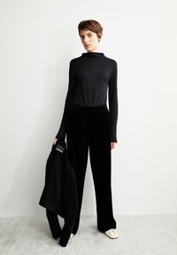 Schwarzes Langarmoberteil mit hohem Kragen, kombiniert mit schwarzen Samenhosen. Das Model hält eine schwarze Jacke mit einem Etikett. Helle Schuhe runden das Outfit ab.