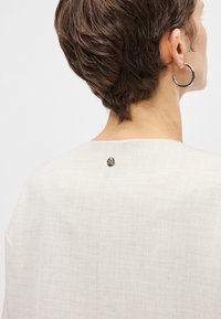 Ανοιχτό μπεζ τοπ με καθαρό, μινιμαλιστικό σχέδιο, που διαθέτει στρογγυλή λαιμόκοψη και μια μικρή στρογγυλή μεταλλική λεπτομέρεια στο πίσω μέρος.