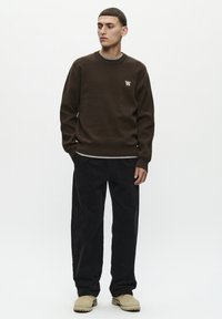 Brauner Strickpullover mit kleinem Logo auf der Brust, kombiniert mit schwarzen Cordhosen und beigen Slippern. Einfacher, lässiger Schnitt.