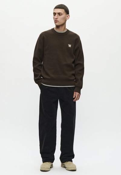 Sudadera de punto marrón con un pequeño logo en el pecho, combinada con pantalones de pana negros y zapatos slip-on beige. Ajuste simple y relajado.
