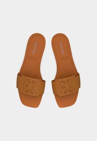 Paire de sandales à slide marron Calvin Klein avec détail de logo découpé sur les brides, semelle plate et design à bout carré sur fond blanc.