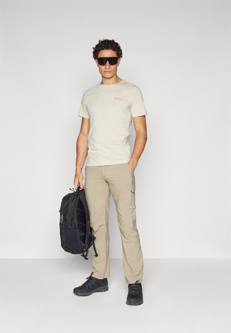 T-shirt beige avec petit logo, pantalon cargo beige avec poches, et chaussures de randonnée noires. Tenant un sac à dos noir. Lunettes de soleil noires.