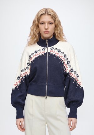Un mannequin blond pose devant un fond uni, vêtu d’une veste brodée à fermeture éclair bleu marine et crème, d’un pantalon crème et de chaussures à talons bleues. Il porte un sac à main bleu marine.