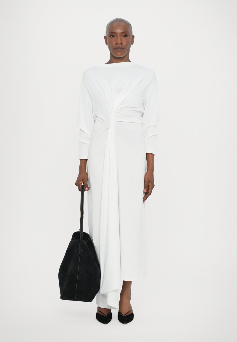 Witte jurk met lange mouwen en een gedraaide voorkant, gemaakt van soepel materiaal. Gecombineerd met een zwarte suède tas en zwarte puntige ballerina's.