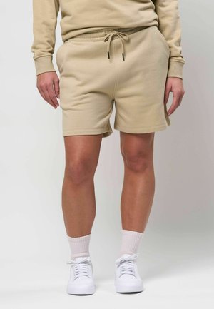 Beine, die beige Shorts mit Kordelzug, weiße Mid-Calf-Socken und weiße Turnschuhe tragen, mit entspannt an den Seiten hängenden Händen vor neutralem Hintergrund.