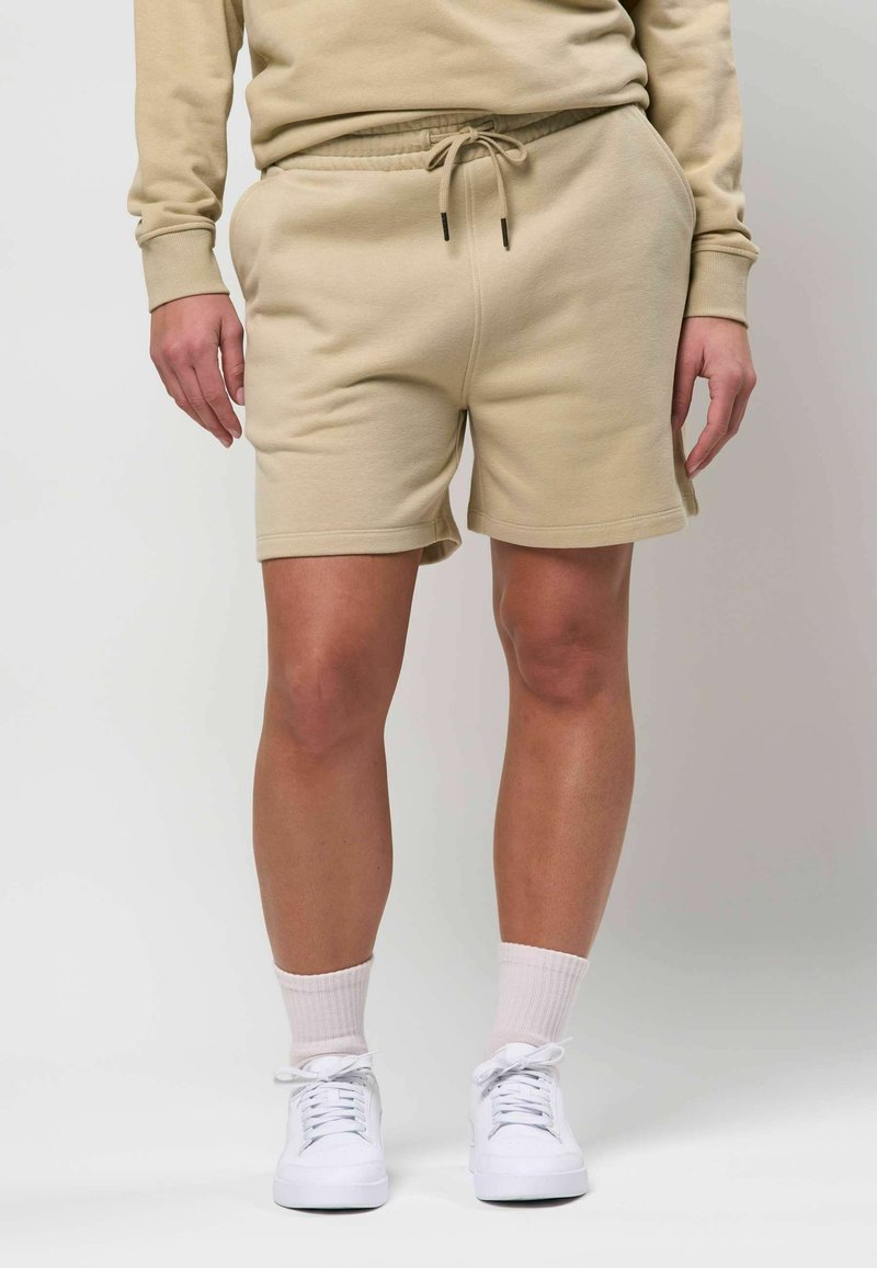 Jambes portant un short beige à cordon, des chaussettes blanches montantes et des baskets blanches, les mains relâchées le long du corps sur un fond neutre.