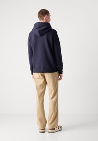Felpa blu navy con cappuccio regolabile, polsini e orlo a coste, abbinata a chinos beige chiaro e sandali marroni e bianchi. Design minimal.